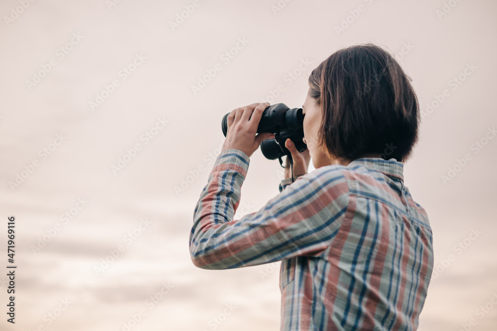 Sticker young unrecognizable woman looks through binoculars during the sunset in the background the sky.