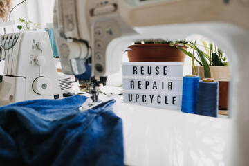 Reuse, repair, upcycle text on light board on sewing machines background. Stack of old jeans, Denim clothes, scissors, thread and sewing tools in sewing studio. Denim Upcycling Ideas, Using Old Jeans - obrazy, fototapety, plakaty
