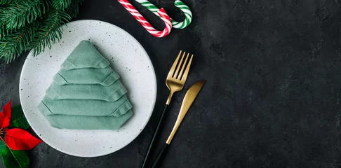  Christmas Dinner Table Setting with empty plate, golden cutlery, green xmas tree napkin on dark background. © nblxer