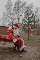 sad man dressed as Santa Claus with tangerines in a package sits on a children's slide. The environmental problem of warming when there is no snow in winter.