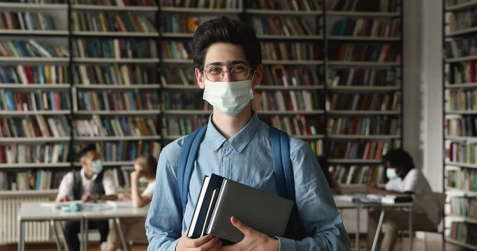 Student guy wear eyeglasses and protective medical face mask holding textbooks, posing standing in university library looking at camera. Studies during corona virus pandemic outbreak, safety concept