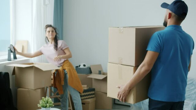 Male mover wearing uniform is bringing cardboard boxes to woman's new apartment helping with relocation. Delivery service and accommodation concept.
