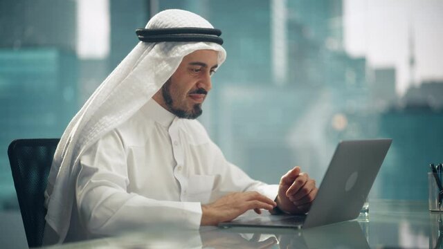 Arab Businessman in White Traditional Outfit Sitting in Office and Working on Laptop Computer. Business Manager Celebrates Successful Investment Deal. Saudi, Emirati, Arab Businessman Concept.