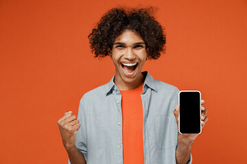 Young black man 50s wearing blue shirt t-shirt hold in hand use mobile cell phone with blank screen workspace area do winner gesture isolated on plain orange color background People lifestyle concept