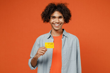 Young smiling rich happy student black man 50s wearing blue shirt t-shirt look camera hold in hand credit bank card isolated on plain orange color background studio portrait. People lifestyle concept.