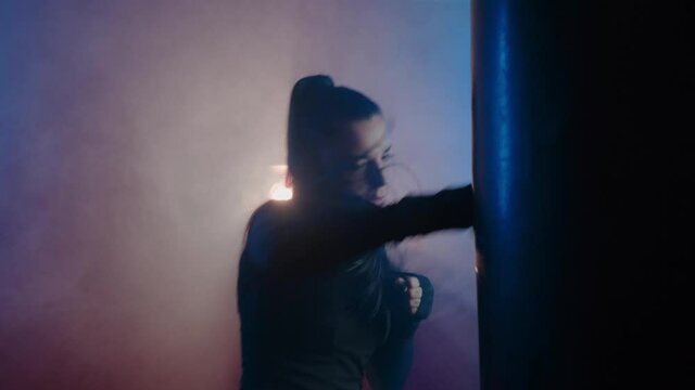 Beautiful strong woman kickboxer alone is engaged in a dark sports hall, beats a punching bag, in slow motion. Aggressive sports woman training endurance strong workout. High quality 4k footage