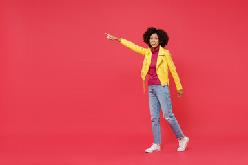 Full size body length happy fancy young curly black latin woman 20s years old wear yellow jacket go move pointing on workspace area copy space mock up isolated on plain red background studio portrait