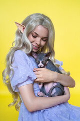 Young female cosplay elf in blue dress smiles happily, gently hugging Sphinx kitten to her chest and looking down at it. Elf has gorgeous blonde curly long hair, pearls in ear. Yellow background.