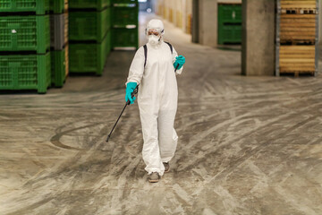The serious situation with the coronavirus. Suppression of COVID19 and cleaning place in factory warehouse by a person in a protective white suit. Cleaning sprayer, stay protective, corona situation
