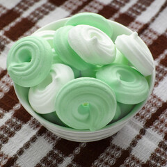 Colored meringue cakes in a white bowl on a checkered kitchen napkin. Top view, flat lay.