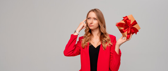 Fototapeta premium Beautiful young woman holding gift box decorated red bow and thinking what to give a friend for birthday, standing over gray background with copy free space for text
