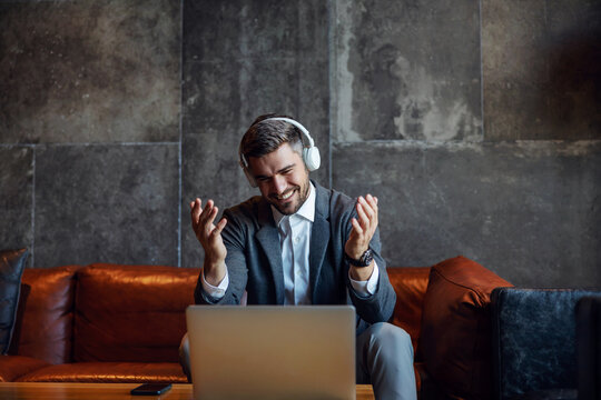 Happy Businessman Wearing Headphones And Sitting In A Hall Of A Hotel And Having A Conference Call With Business Partners. They Have An Agreement. Telecommunications, Online Meeting