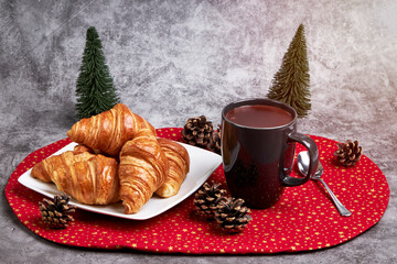 Cup of hot chocolate with a plate with croissant on a red Christmas tablecloth and some Christmas fir trees