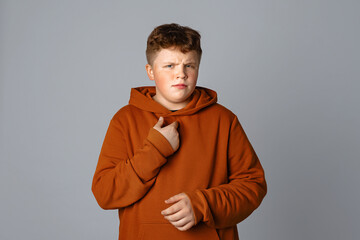 Indignant displeased plus size teenager boy pointing hand at himself, standing over gray background, having doubtful confused uncertain facial expression
