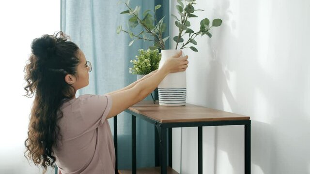 Young woman is decorating house with flowers putting plants in pots and vases on shelves busy with housework during relocation. Youth and domestic chores concept.