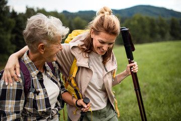 Happy mid adult woman with trekking poles hiking with active senior mother outdoors in nature.