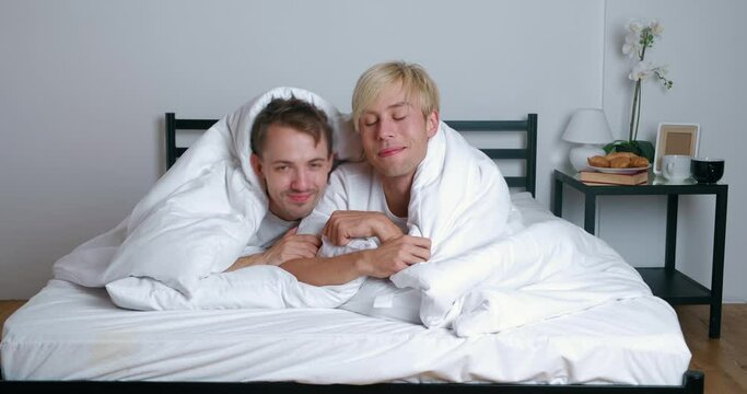 Young gay couple lying on bed together in bedroom, hugging and kissing. LGBT family