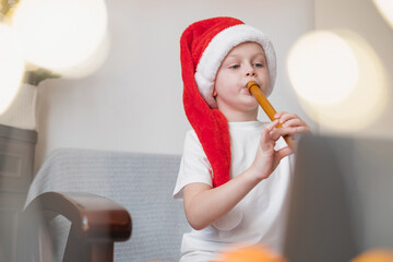 Boy playing wind musical instrument at home.child wearing Santa hat. On background gold Christmas lights. Online training, online classes.