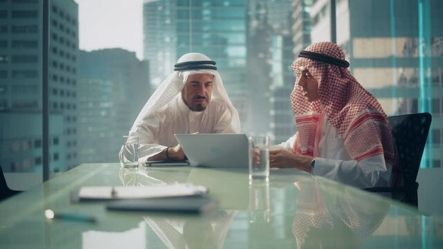 Two Successful Emirati Businessmen in White Traditional Kandura Sitting in Office Meeting, Negotiating and Talking About Investment Opportunities, Using Laptop. Saudi Emirati, Arab Businessman Concept