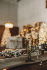 chic Christmas table setting in beige and wooden colors, glassware with gold cutlery, candles, cotton and garlands against a bokeh background and Christmas interior, selective focus