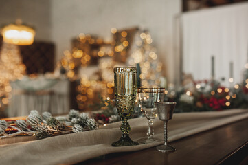glass on background of chic Christmas table setting in beige and wooden tones, glassware with gold cutlery, candles, cotton and garlands on the background of bokeh and Christmas interior