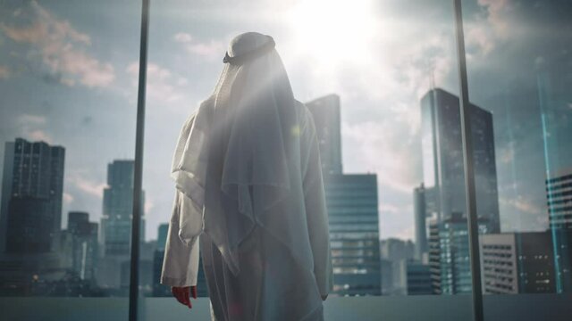 Successful Muslim Businessman in Traditional White Outfit Walking in His Modern Office Looking out of the Window on Big City with Skyscrapers. Successful Saudi, Emirati, Arab Businessman Concept.