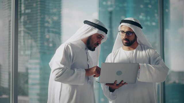 Two Successful Muslim Businessmen in White Traditional Outfit Standing in Office Meeting, Negotiating and Talking About Financial Opportunities, Using Laptop. Saudi, Emirati, Arab Businessman Concept.