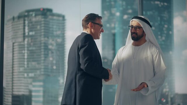 International Operations Manager Meeting Muslim Business Partner in Traditional White Kandura. They're Standing in Modern Office, Shaking Hands. Successful Saudi, Emirati, Arab Businessman Concept.
