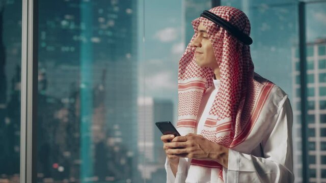 Young Muslim Businessman in Traditional White Kandura Standing in His Modern Office, Using Smartphone Next to Window with Skyscrapers. Successful Saudi, Emirati, Arab Businessman Concept.