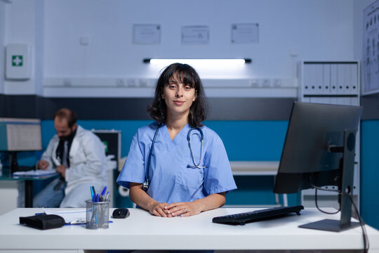 Portrait Of Medical Assistant Using Computer At Desk For Work Overtime. Woman Nurse In Uniform Looking At Camera Preparing For Paperwork And Healthcare Checkup, Working Late At Office.