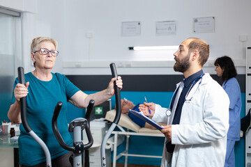 Old patient doing physical exercise with stationary bicycle while doctor doing consultation for physiotherapy. Retired woman using bike for fitness workout to recover from muscle injury