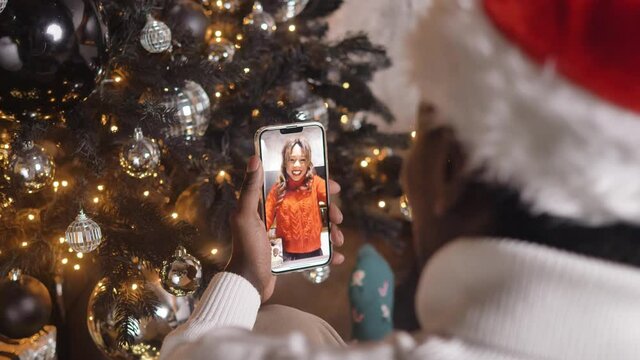 African American man in Santa hat and knitted sweater talks to white girlfriend via videocall using smartphone against Christmas tree