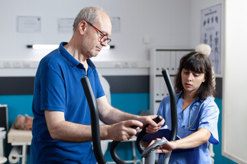 Retired patient doing physical exercise for injury recovery with medical assistant. Man using stationary bicycle for aerobics and gymnastics to treat muscle pain with physiotherapy.