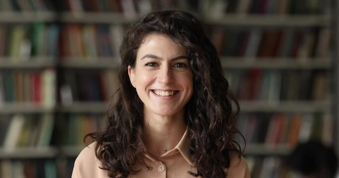 Close up attractive brunette teenager girl with curly hair and charming smiling posing in library look at camera. Head shot of higher education institution student portrait, excellent studies concept