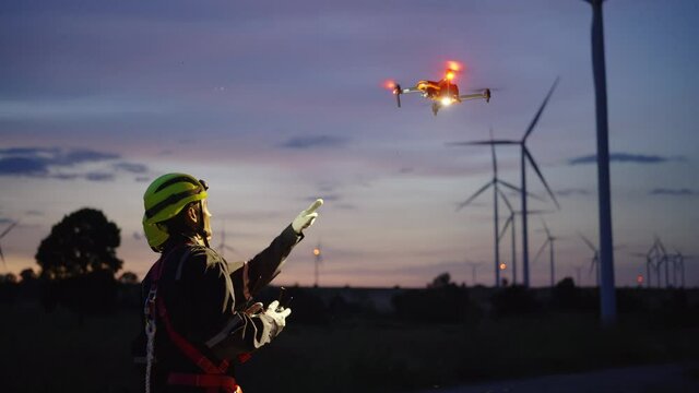 Two Asian technicians in a safety kit fly the drone to inspect the wind turbine's operation.