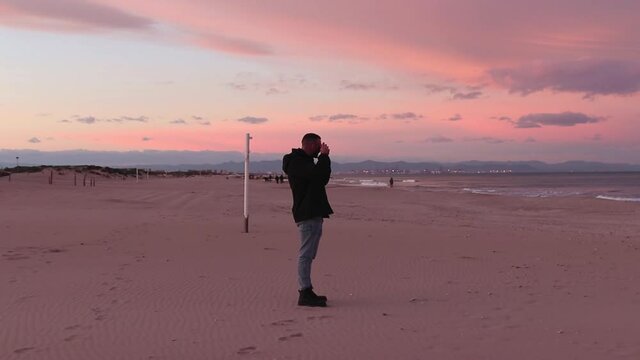 Man with black jacket taking a picture with his analog camera on a beach at sunset in winter