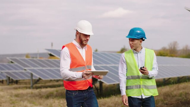 Concept of electrical innovation two multiracial ecological workers with safety equipment walking in front of the camera through the solar panels power station and discussing something
