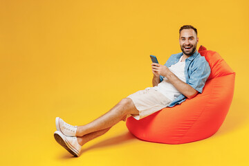 Full body young smiling happy fun amazed man 20s wearing blue shirt white t-shirt sit in bag chair use mobile cell phone isolated on plain yellow background studio portrait. People lifestyle concept