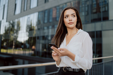 Attractive girl working in office corporation, company, dressed in elegant clothes, stands leaning against railing in front of glass building, holding phone in hands, waiting for call, looking to side