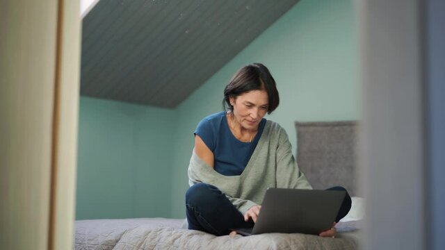 Positive mature woman typing by laptop on the bed