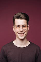 Isolated shot of young handsome male with glasses trendy hairdo, wears casual brown t shirt, has serious expression as listens to interlocutor, poses in studio against crimson background