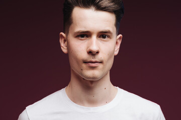 Isolated shot of young serious handsome male with trendy hairdo, wears casual white t shirt, has serious expression as listens to interlocutor, poses in studio against crimson background