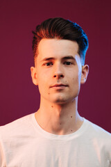 Isolated shot of young serious handsome male with trendy hairdo, wears casual white t shirt, has serious expression as listens to interlocutor, poses in studio against crimson background
