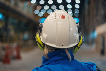 Back view of male worker wearing work vest and safety helmet while looking at industrial equipment...