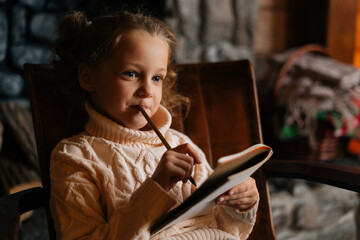 Close-up face of smiling thoughtful little blonde curly child girl writing letter to Santa Claus sitting on chair at home with festive interior. Pretty Caucasian kid writes letter with wishes for gift