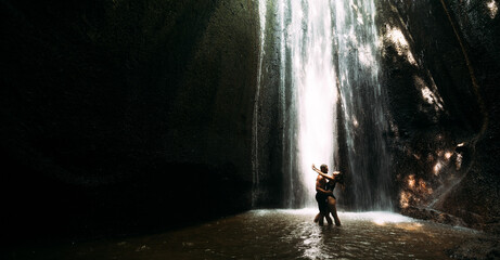 Beautiful couple in a cave with a waterfall. Athletic man and woman under the streams of a waterfall. A sexy couple under a tropical waterfall in twilight light. Tukad Chepung Waterfall. Copy space