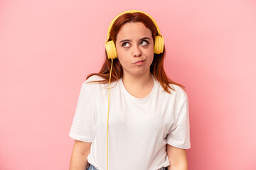 Young caucasian woman listening to music isolated on pink background confused, feels doubtful and unsure.