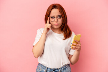 Young caucasian woman holding a mobile phone isolated on pink background pointing temple with finger, thinking, focused on a task.