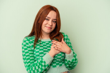 Young caucasian woman isolated on green background has friendly expression, pressing palm to chest. Love concept.