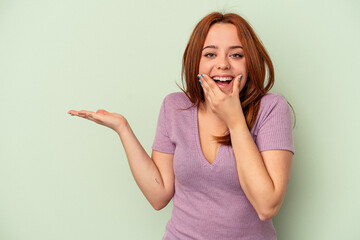 Young caucasian woman isolated on green background holds copy space on a palm, keep hand over cheek. Amazed and delighted.